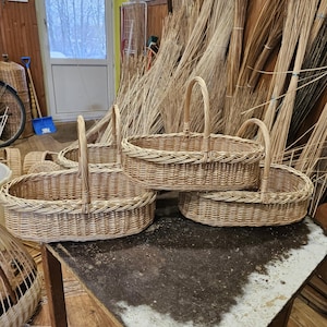 May include: Several woven wicker baskets with arched handles sit on a weathered table. The oval-shaped baskets are light brown and appear to be handmade. The background shows more wicker materials and a window.
