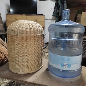 Può includere: Un cesto in vimini intrecciato marrone chiaro con coperchio arrotondato è accanto a un grande contenitore d'acqua blu. Il cesto è cilindrico. Il contenitore è trasparente con base blu ed etichetta bianca.
