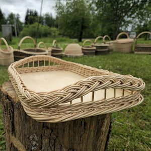 Puede incluir: Una cesta rectangular de mimbre con base de madera clara y borde trenzado. La cesta está sobre un tocón de árbol, con otras cestas de mimbre al fondo. La cesta está hecha de material tejido marrón claro.