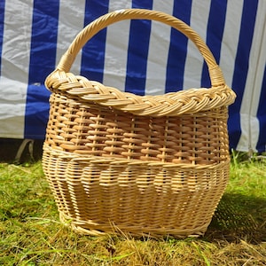 Può includere: Un cesto in vimini intrecciato, di forma arrotondata e con un manico arcuato. Il cesto è realizzato in vimini marrone chiaro e beige, con un bordo intrecciato. È appoggiato su erba verde, con uno sfondo a righe blu e bianche.