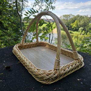 Puede incluir: Una cesta rectangular de mimbre trenzado con una base de madera de color tostado claro y un asa arqueada única. La cesta presenta un borde trenzado a lo largo de los lados y está construida con varas de sauce natural de color claro.