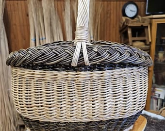 May include: An oval-shaped woven basket featuring a light beige centre section, dark brown bands at the top and bottom, and a braided rim. The basket includes a single upright handle made of light-coloured woven material attached to the centre.