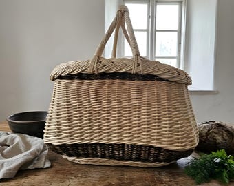May include: A large, oval-shaped woven basket featuring a light beige wicker body with a dark brown woven base and a matching lid. The basket includes two upright handles that meet at the top, with a braided rim detail around the lid edge.