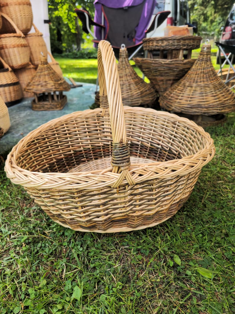 May include: A large, oval-shaped woven basket with a tall handle. The basket is made of light brown and dark brown wicker, creating a textured pattern. The handle is tightly woven and curves upwards. Other woven baskets and a birdhouse are visible in the background.