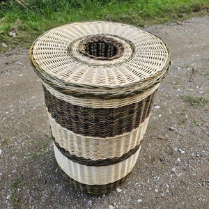 Puede incluir: Una cesta cilíndrica de mimbre trenzado con bandas horizontales alternas de fibras naturales en color crema claro y marrón oscuro. La cesta incluye una tapa circular a juego con una abertura central y un borde trenzado de color marrón oscuro.