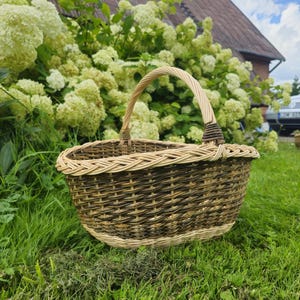 May include: Oval-shaped woven basket with a handle. The basket is made of natural and dark brown wicker, with a light-colored rim and handle. The basket is sitting on green grass.