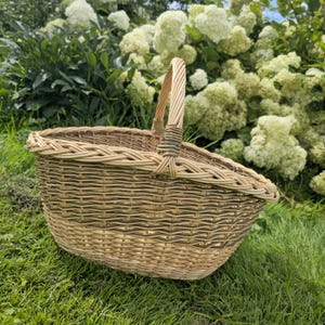 May include: A woven wicker basket with a curved handle. The basket is oval-shaped and has a natural, light brown color with green accents. It sits on a bed of green grass, with white flowers in the background.
