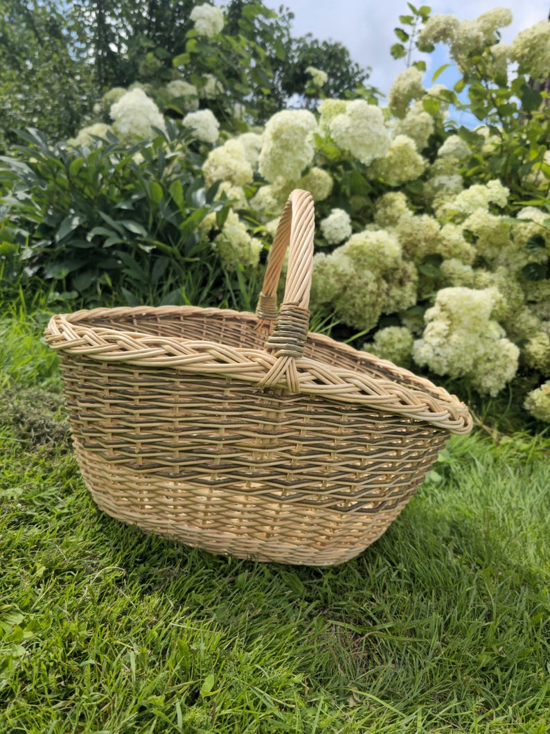 May include: A woven wicker basket with a curved handle. The basket is oval-shaped and has a natural, light brown color with green accents. It sits on a bed of green grass, with white flowers in the background.