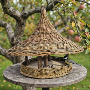 May include: A round bird feeder constructed from woven brown willow branches featuring a wide, conical roof and a flat circular base. The structure is supported by several vertical wooden posts, and a small yellow and black bird sits inside.