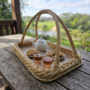 May include: A rectangular woven wicker serving tray with a high arched handle. The tray holds a white ceramic teapot, two clear glass cups filled with amber tea, and two small plates containing chocolate chip cookies.