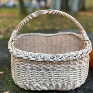 Può includere: Un cestino in vimini intrecciato beige chiaro con manico curvo. Il cestino ha una forma rettangolare con un bordo intrecciato attorno al bordo superiore. Il manico è anch'esso intrecciato e attaccato ai lati del cestino.