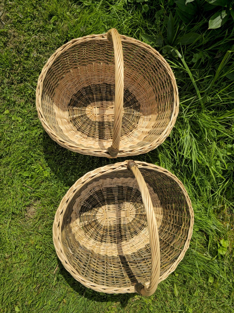 May include: Two woven wicker baskets with handles. The baskets are oval-shaped and have a natural, light brown color with darker brown accents in the center. The handles are arched and made of the same woven material. The baskets are placed on a bed of green grass.