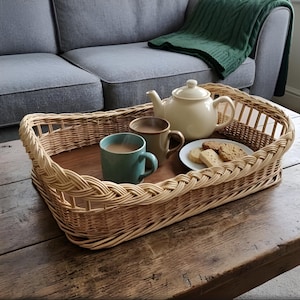 Puede incluir: Una bandeja rectangular de mimbre trenzado con base de madera oscura contiene una tetera de cerámica color crema, una taza verde azulado, una taza marrón y un plato blanco pequeño con galletas, sobre una superficie de madera.