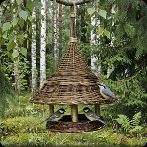 Op de afbeelding: Een hangende vogelvoederbak gemaakt van gevlochten bruine wilgentakken, met een kegelvormig dak en een ronde basis. Twee geel-zwarte koolmezen en een grijs-witte boomklever zitten op de structuur, die aan een koord is opgehangen.