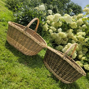 May include: Two woven wicker baskets with arched handles rest on green grass. The baskets have a natural brown and tan color scheme, with a lighter border around the top. The background features white flowers and lush greenery.