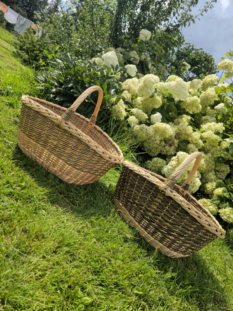 May include: Two woven wicker baskets with arched handles rest on green grass. The baskets have a natural brown and tan color scheme, with a lighter border around the top. The background features white flowers and lush greenery.