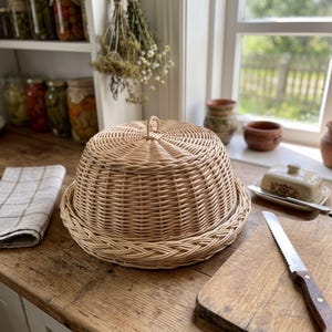 May include: A round, light brown woven wicker food cover with a dome shape and a small loop handle at the top. The cover features a braided border along the base and a tight, circular weave pattern throughout the structure.
