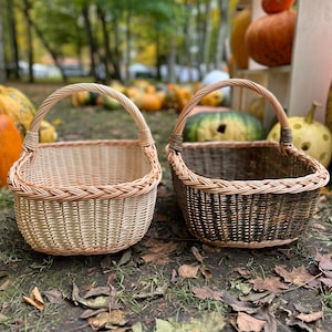 May include: Two woven wicker baskets with arched handles. One basket is light tan, the other is dark brown. The baskets are sitting on the ground with fallen leaves, with pumpkins in the background.