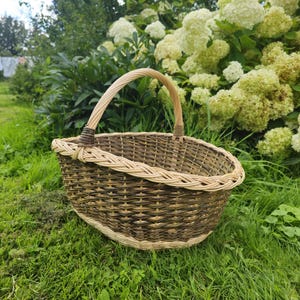 May include: A woven basket with a handle, resting on green grass. The basket is made of brown and tan wicker, with a rounded shape and a sturdy handle. The background features lush greenery and white flowers.