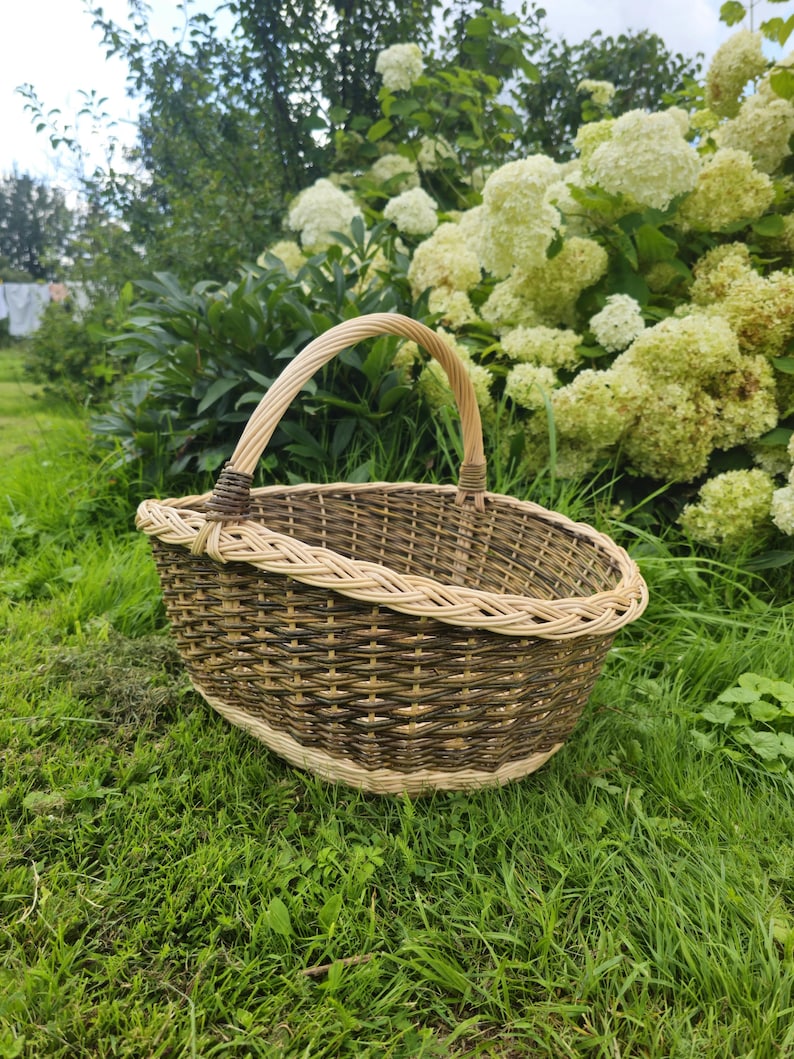 May include: A woven basket with a handle, resting on green grass. The basket is made of brown and tan wicker, with a rounded shape and a sturdy handle. The background features lush greenery and white flowers.