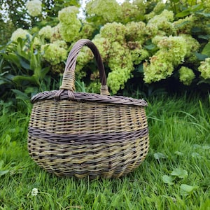 May include: A round, handwoven wicker basket with a handle, set on green grass. The basket has alternating bands of light and dark brown, with a strong handle. The background features lush green foliage and white flowers.