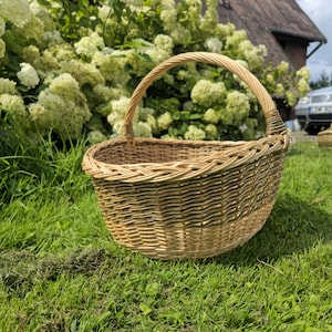 May include: A round, woven wicker basket with a sturdy handle. The basket is made of light brown and tan wicker, and sits on a bed of green grass. The handle is arched and woven for a comfortable grip. The basket is designed for carrying items.
