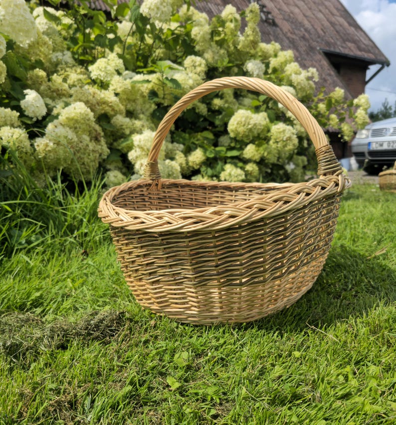 May include: A round, woven wicker basket with a sturdy handle. The basket is made of light brown and tan wicker, and sits on a bed of green grass. The handle is arched and woven for a comfortable grip. The basket is designed for carrying items.
