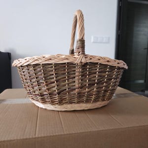 May include: A woven wicker basket with a handle. The basket is oval-shaped with a light brown rim and a darker brown body. The handle is made of twisted wicker. The basket is sitting on a cardboard box.