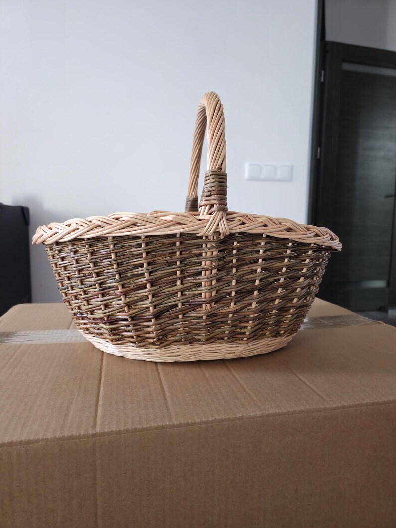 May include: A woven wicker basket with a handle. The basket is oval-shaped with a light brown rim and a darker brown body. The handle is made of twisted wicker. The basket is sitting on a cardboard box.
