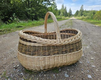 May include: An oval-shaped woven basket featuring a two-tone design with a light beige base and a darker brown upper section. The basket includes a single arched handle and a thick, braided rim detail crafted from natural willow reeds.
