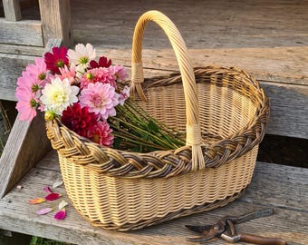May include: A woven wicker basket with a single arched handle, featuring a braided rim design. Inside the basket is a bouquet of pink, white, and deep red dahlia flowers. A pair of metal garden shears lies next to the basket on the wooden surface.
