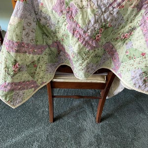 May include: A patchwork quilt featuring a repeating geometric pattern of triangles and squares. The fabric displays a mix of light green, lavender, and white colors with floral prints, polka dots, and gingham checks, finished with a cream-colored border.