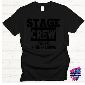 May include: Black t-shirt with the words "STAGE CREW" above "I WORK IN THE SHADOWS" in black block letters. The shirt has a crew neck and short sleeves. The shirt is a Gildan brand.