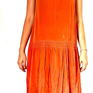 May include: A sleeveless, orange dress with a V-neck and a gathered waist. The dress has a flowing skirt and is made of a soft, silky material. The dress is a vibrant shade of orange and appears to be vintage.