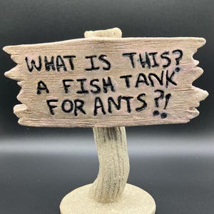 May include: A wooden sign with the text "What is this? A fish tank for ants?!" on a brown, textured base.