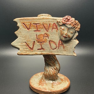 May include: A ceramic sculpture of a sign with the words "Viva la Vida" written on it. The sign is held up by a twisted tree trunk and has a small ceramic face with a flower crown on the side.
