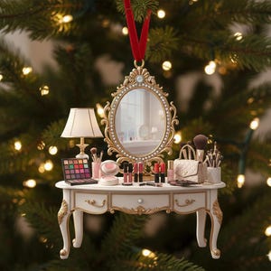 May include: A festive Christmas tree ornament depicting a miniature vanity table. The white table features gold detailing and is decorated with tiny makeup products, a mirror, and a lamp. It hangs from a red ribbon, set against a backdrop of Christmas tree lights.