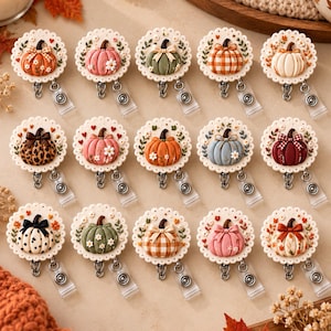 May include: A collection of fifteen circular badge reels, each featuring a unique pumpkin design on a scalloped white base. The pumpkins vary in color, pattern, and decorative accents, including bows, flowers, and leaves, attached to retractable clips.