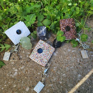 May include: Glitter badge reels in white, brown, and multicoloured glitter, each with a black button and a silver keychain. The reels are displayed on a concrete surface with green foliage in the background.