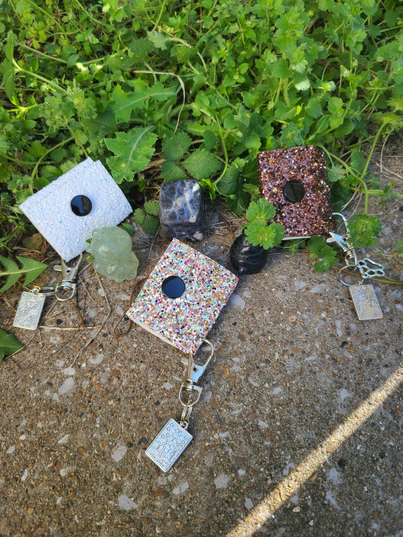 May include: Glitter badge reels in white, brown, and multicoloured glitter, each with a black button and a silver keychain. The reels are displayed on a concrete surface with green foliage in the background.