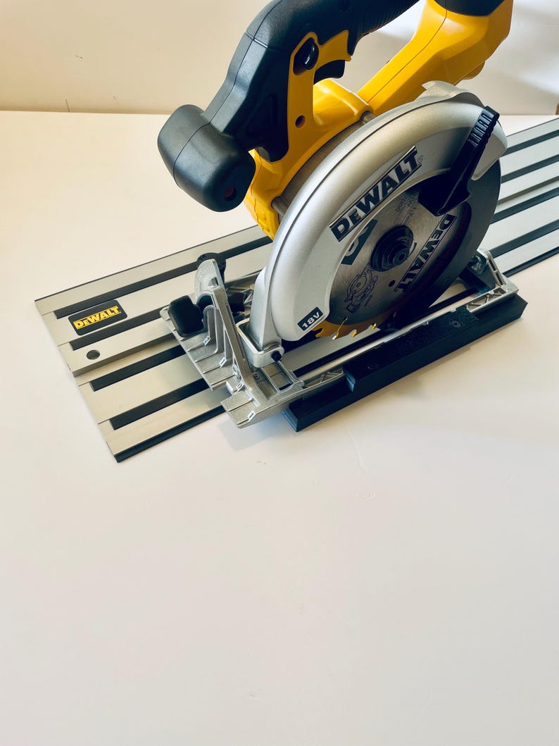 May include: A yellow and black DEWALT circular saw with a silver blade and a black guide rail. The saw is resting on a white surface. The saw blade is 18V.