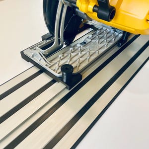 May include: A yellow and black circular saw with a silver base is positioned on a white and black track system. The saw features a black blade guard and handle. The track system has alternating white and black stripes.