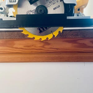 May include: A yellow and silver circular saw blade with a black guard is mounted on a white and yellow power tool. The blade is 165mm in diameter and has the text "NMAX 7000" and "18V" printed on it. The saw is positioned over a wooden surface.