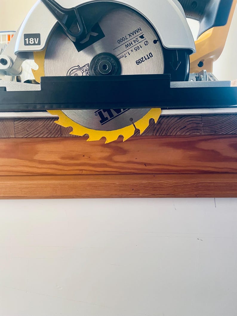 May include: A yellow and silver circular saw blade with a black guard is mounted on a white and yellow power tool. The blade is 165mm in diameter and has the text "NMAX 7000" and "18V" printed on it. The saw is positioned over a wooden surface.