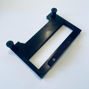 May include: A black rectangular frame with two cylindrical posts and a rectangular opening. The frame has a textured surface and is set against a white background. The frame appears to be made of plastic.