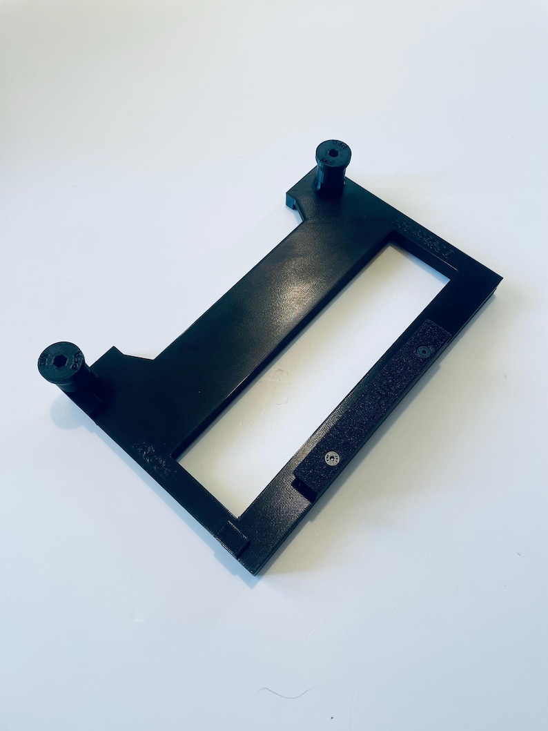 May include: A black rectangular frame with two cylindrical posts and a rectangular opening. The frame has a textured surface and is set against a white background. The frame appears to be made of plastic.