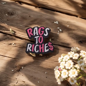 May include: Embroidered patch with the text "Rags to Riches" in a stylized font. The patch features a black border, white lettering, and pink accents, including a dollar sign. The patch is set against a wooden background.