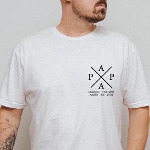 Può includere: Una t-shirt bianca a maniche corte con una grafica nera sul petto sinistro. Il design presenta la parola PAPA disposta a croce, con i nomi Charlotte e Amelie e gli anni EST. 2018 e EST. 2022 stampati al di sotto.