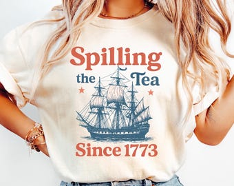May include: Cream-colored t-shirt featuring a vintage navy blue sailing ship graphic. The text "Spilling the Tea Since 1773" is printed in red and navy blue. The shirt has rolled-up sleeves and is paired with blue jeans.
