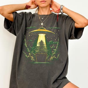 May include: A charcoal grey short-sleeve t-shirt featuring a graphic print of a yellow UFO hovering over a green potted plant. The design includes a bright yellow light beam shining down from the saucer onto the plant, surrounded by green foliage.